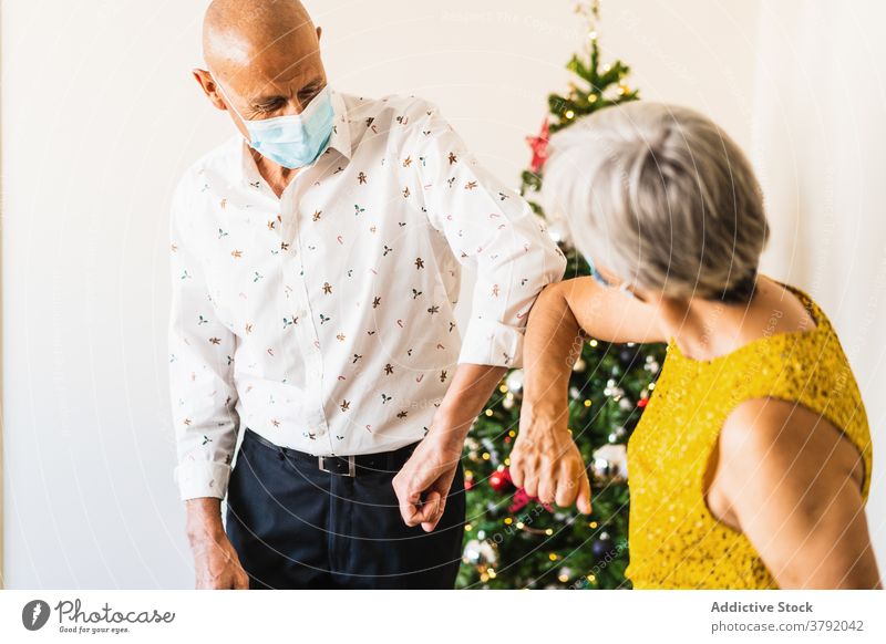 Mature couple in masks bumping with elbows near Christmas tree greet christmas festive eve coronavirus new normal salute positive mature greeting cheerful