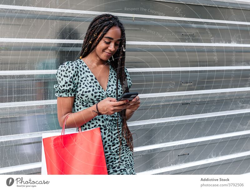 Smiling black woman using smartphone on street shopping bag paper bag shopaholic style trendy summer female ethnic african american dress city metal wall