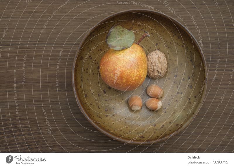 Autumnal composition on dark oak wood from brown ceramic bowl with pear walnut and three hazelnuts Text space Pear fruit Walnut Monochrome Leaf Brown Green