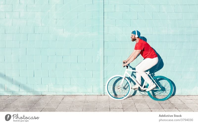 Young man riding bicycle and listening to music in headphones on city street ride bike cyclist active trick trendy building stone wall male young beard outfit