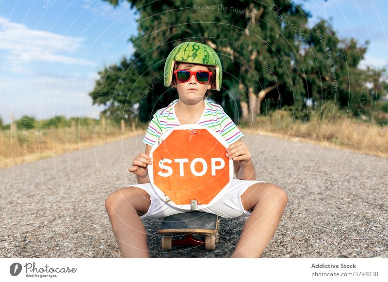 Serious boy in decorative head wear with signboard on roadway stop prohibit restriction trendy headwear sunglasses skateboard show watermelon kid summer style