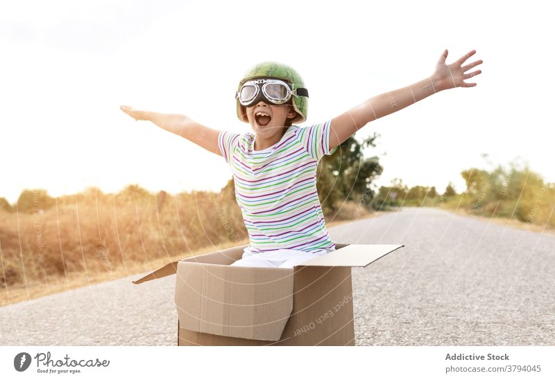 Excited boy in goggles sitting in cardboard box on road excited having fun childhood mouth opened stylish carefree sky arms raised kid positive smile enjoy