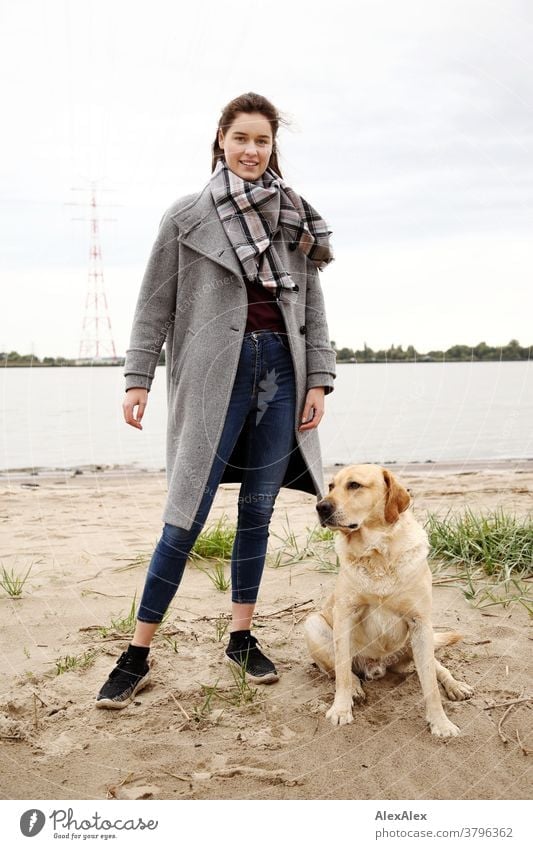 Close portrait of a young woman at the Elbe beach with blond Labrador Woman Young woman 18-25 years warmly pretty Charming Slim Brunette long hairs Fresh Large