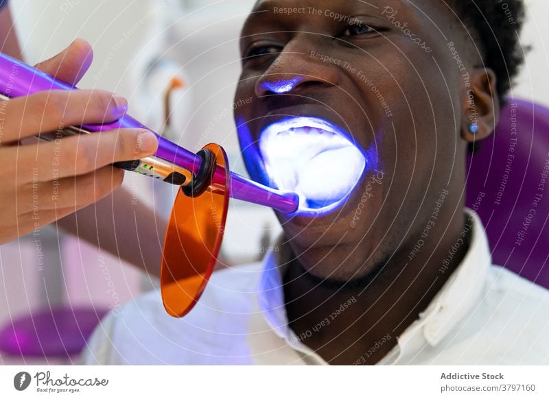 Crop doctor using dental curing light during procedure cure dentist patient stomatology uv ultraviolet treat ethnic black african american clinic medical chair