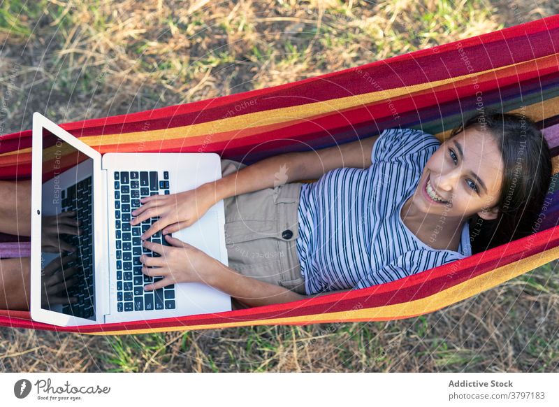 Smiling woman in hammock with laptop freelance project using typing startup independent cheerful female young internet gadget device netbook browsing lying work