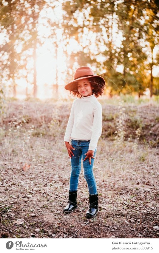 portrait of cute afro kid girl wearing a hat at sunset during golden hour, autumn season, beautiful trees background nature outdoors brown leaves