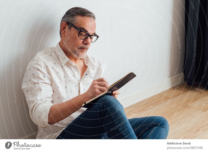 Concentrated man taking notes in notebook while sitting on floor write take note serious focus concentrate diary parquet casual mature at home busy lifestyle