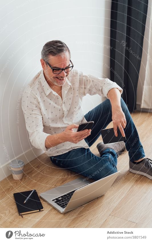 Positive mature man using smartphone and laptop on floor workspace browsing focus positive gadget device watching app concentrate casual at home busy male