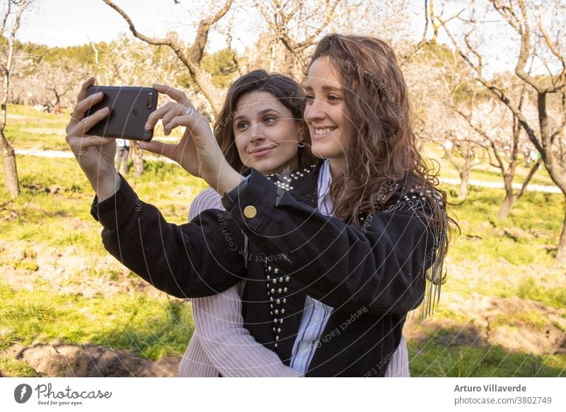 two girls in a park take cell phone selfies while hugging. They are both very pretty and smile happily. One is wearing a pink sweater and the other is wearing a studded jacket