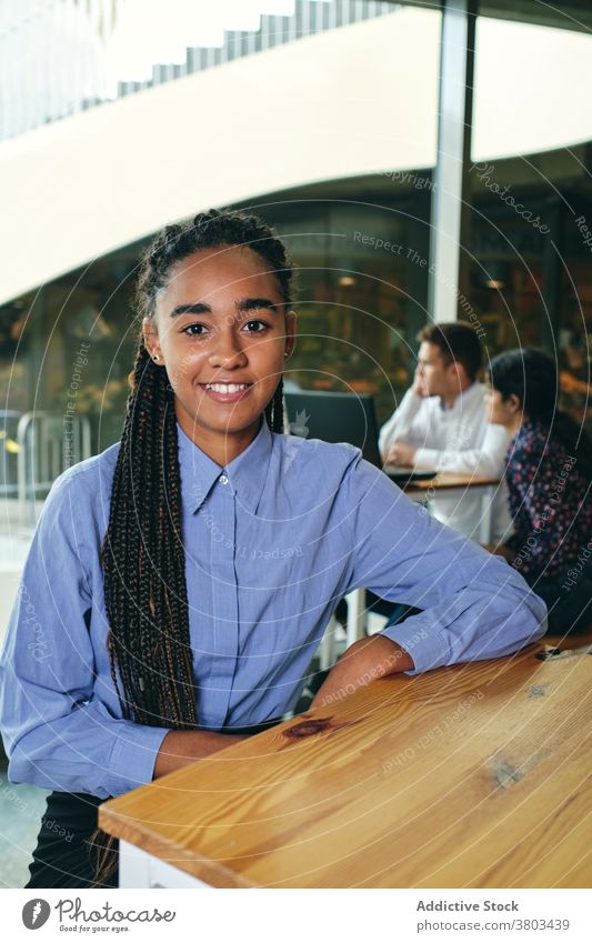 Smiling woman in blue shirt resting at table glad Afro braids confident satisfied sincere friendly pleasant portrait white shirt appearance building smile