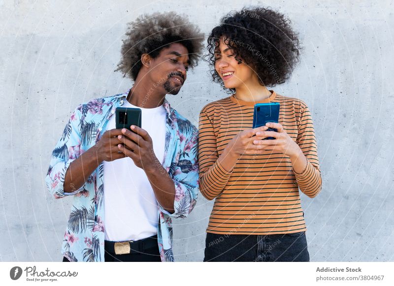 Glad multiethnic friends watching smartphones on street couple content stylish wear hairstyle friendship using gadget cheerful cellphone trendy afro device