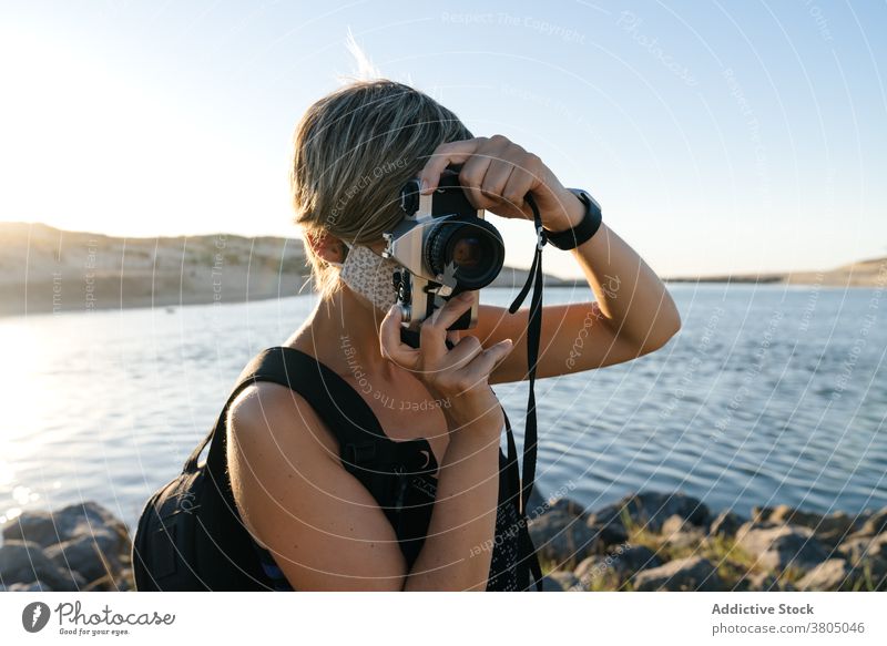 Focused young woman with photo camera relaxing on rocky seashore at sunset coast tourist peaceful admire wanderlust trip holiday vacation female short hair