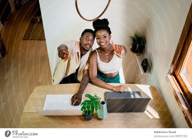 Cheerful young black couple working remotely on laptops at home positive point at camera together relationship cuddle cheerful job busy boyfriend girlfriend
