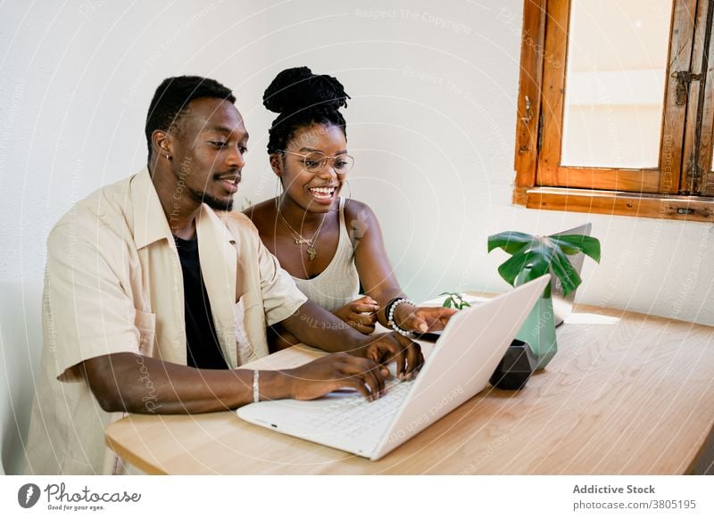 Cheerful young black couple working remotely on laptops at home positive point at camera together relationship cuddle cheerful job busy boyfriend girlfriend