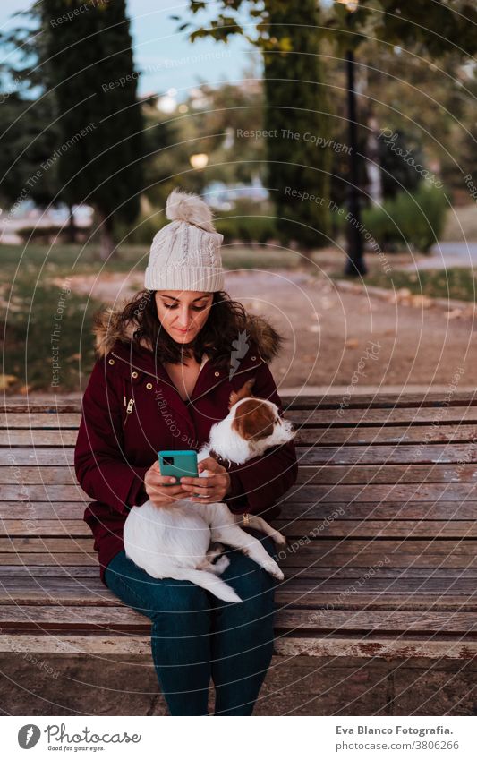 woman sitting on a bench, using mobile phone in a park with her adorable jack russell dog. Lifestyle outdoors autumn brick wall casual clothing caucasian