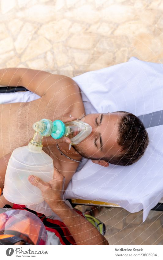 Patient in oxygen mask on ocean shore in summer lifeguard patient aid coworker help covid preoccupied beach medical visor profession emergency protective shield