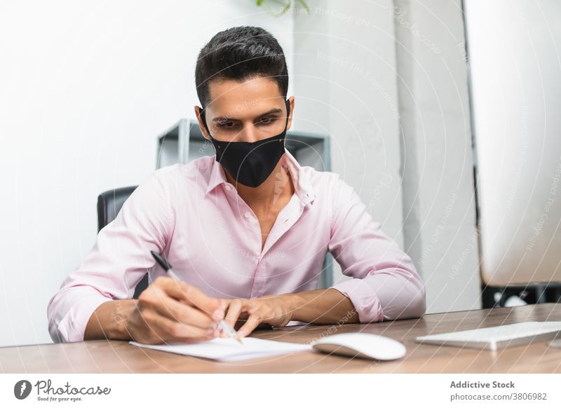 Unrecognizable focused entrepreneur writing on paper near computer in office businessman write work project mask desk take note pen concentrate formal wear