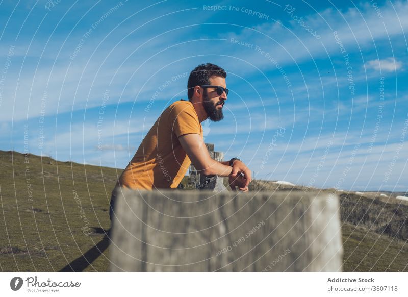 Confident young male traveler resting on grassy hill man relax admire nature confident fence wanderlust calm serene lean on ethnic casual outfit sunglasses