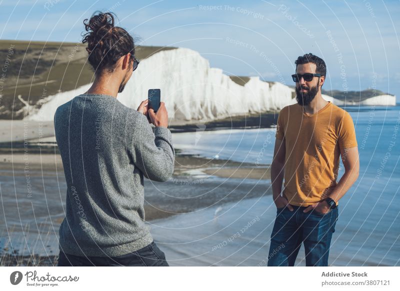 Young man taking photo on smartphone of friend standing on seashore near cliff men take photo spend time holiday friendship nature journey relax vacation young