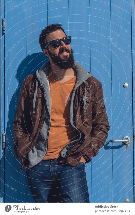 Stylish bearded ethnic man relaxing near cozy house in sunlight hand in pocket style brutal travel door modern confident town chill male young trendy outfit