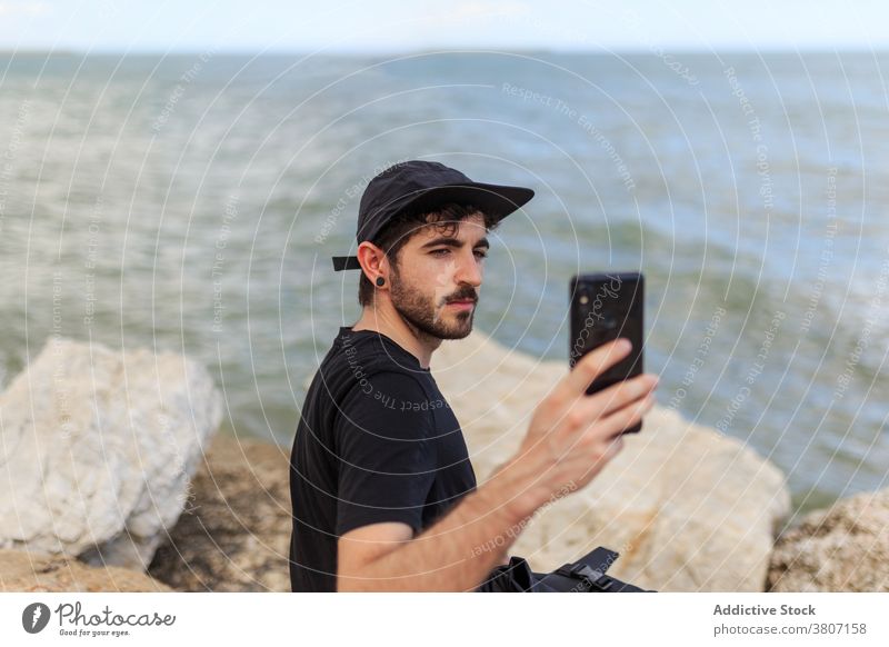 Man surfing internet on smartphone resting on boulders near sea man selfie horizon nature blue sky using device seascape cloudy gadget ocean free time cellphone