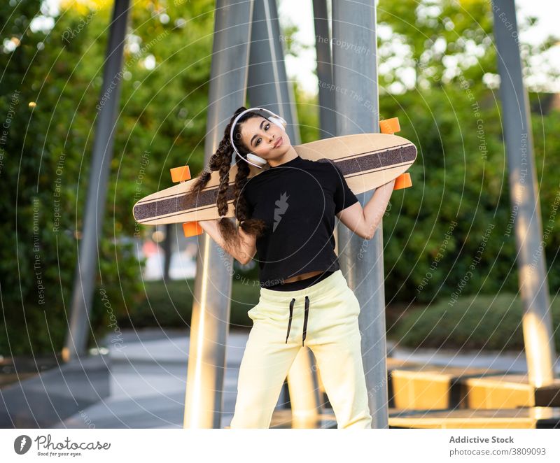 Cheerful ethnic lady with longboard enjoying music in headphones in park woman listen skate park happy active cheerful trendy tree training modern female young