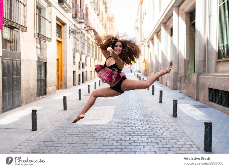 Cheerful young ethnic ballerina jumping on street woman happy dance perform ballet smile flexible energy motion dancer female fit long hair curly hair