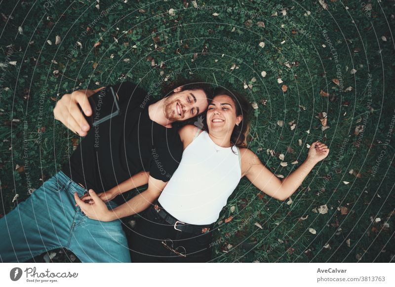 Young couple taking a selfie while lying on the grass people man smart-phone park portrait boy click happy lay down technology oneself nature holding smile girl