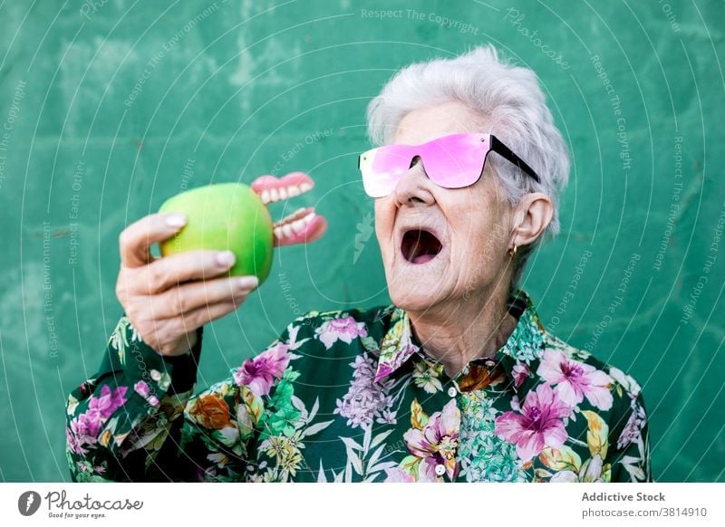 Funny senior woman with apple and denture teeth funny joke humor trendy style bite elderly female sunglasses accessory lady old health care mouth aged retire