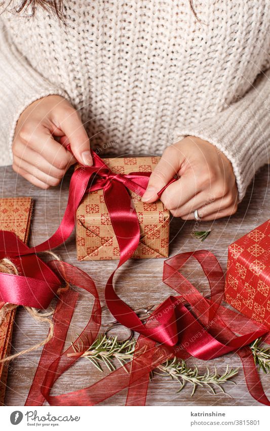 Woman ties a ribbon bow and wrapping presents somebody woman hands paper close up Gift box christmas sweater craft paper red rope rustic table Wrapped Holiday