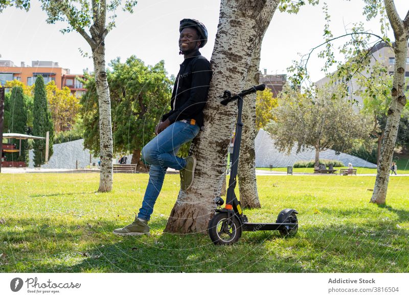Ethnic man near scooter in park electric helmet city urban tree garden protect male ethnic black african american cheerful modern transport smile lean casual