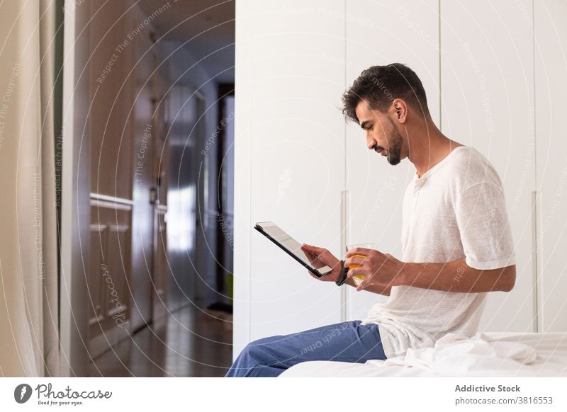 Young man using tablet in bedroom morning drink gadget at home device young beard browsing internet read online lifestyle connection surfing beverage orange