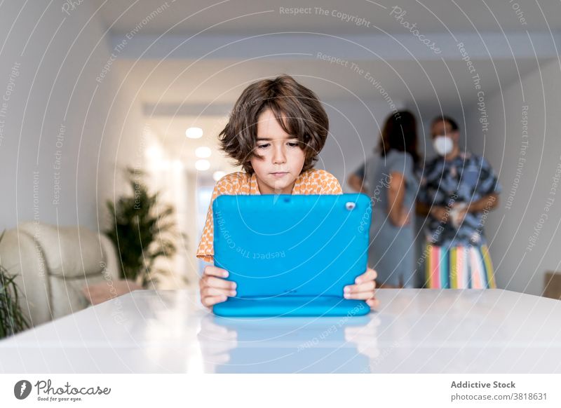 Focused boy watching laptop behind unrecognizable parents at home internet online childhood focus table using gadget device netbook covid mother father mask