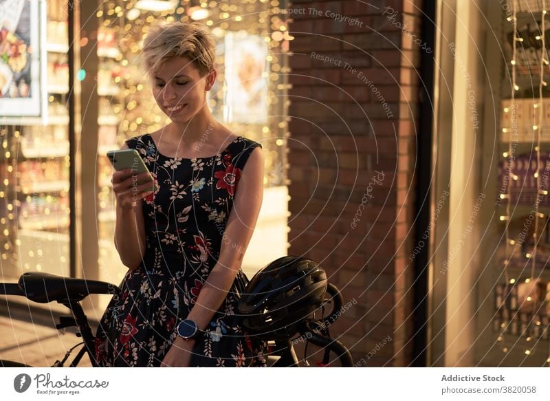 Young woman with phone in hand sits near store and bicycle Woman dress bike evening walk girl young riding beautiful city night lights building glass window