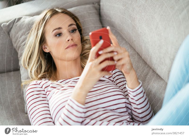 Young blonde woman using her smartphone lying on the sofa. home girl mobile young white cellphone people indoors couch lifestyle living sitting room telephone
