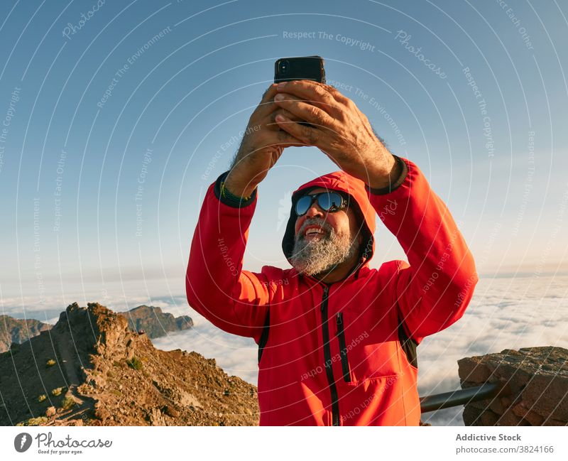 Cheerful elderly traveler taking selfie in mountains man explore hiker mountaineer smartphone senior male outerwear sunglasses smile top sunny summit peak