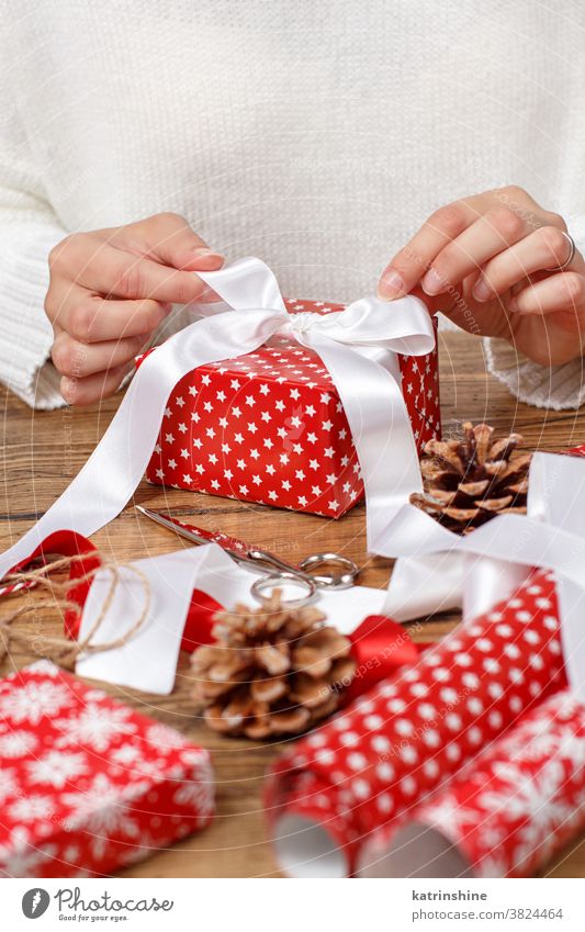 Woman ties a ribbon bow on a wrapped present woman hands close up Gift box christmas sweater red white wooden rustic pune cones table Wrapped Holiday Package