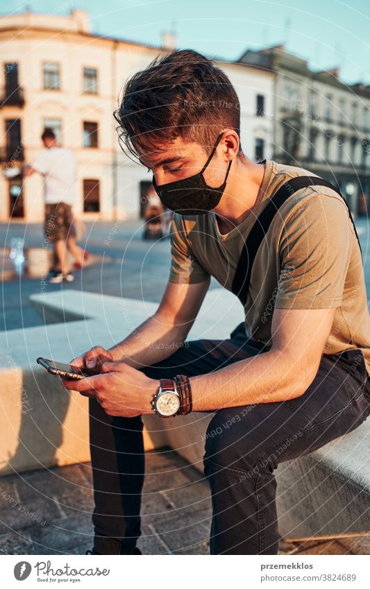 Young man sitting in the city center, using smartphone, wearing the face mask to avoid virus infection caucasian conversation covid-19 lifestyle outbreak
