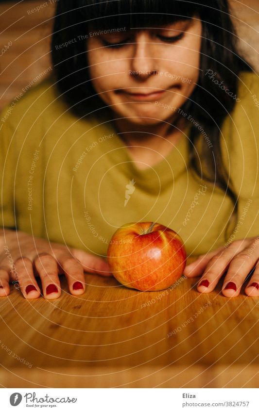 A young woman sits at the table and looks down on an apple in frustration. Diet, healthy food. Healthy Eating Vegan diet Woman Apple frustrated decrease