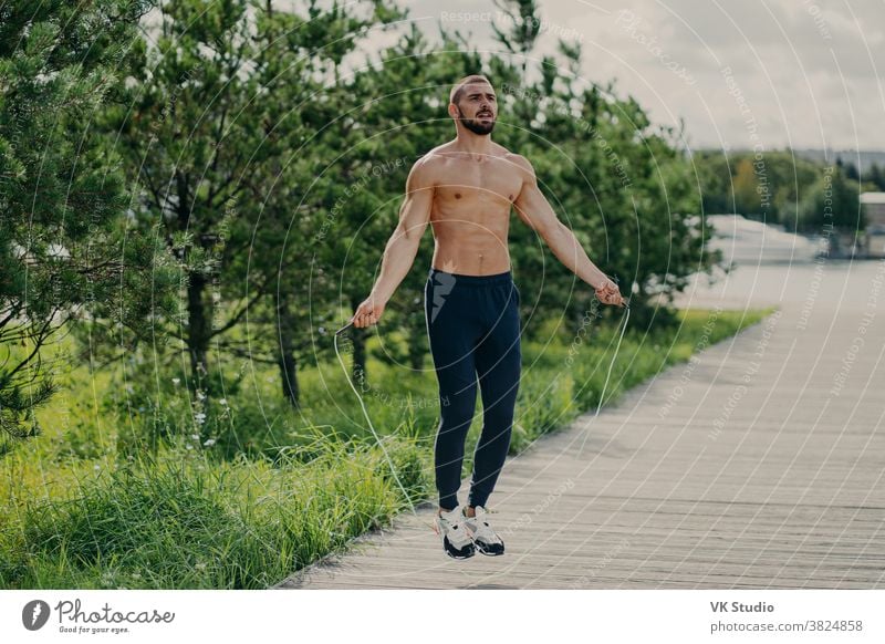 Sporty man with thick beard does exercises with jumping rope, has muscular torso, stays in good physical shape, poses outdoor. Fitness and healthy concept. Male runner warms up with sport equipment
