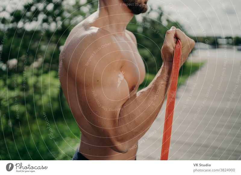 Cropped shot of unrecognizable muscular man has workout biceps exercise stands in profile shows strong muscles, uses resistance band, strives for strong body. Bodybuilding, motivation concept