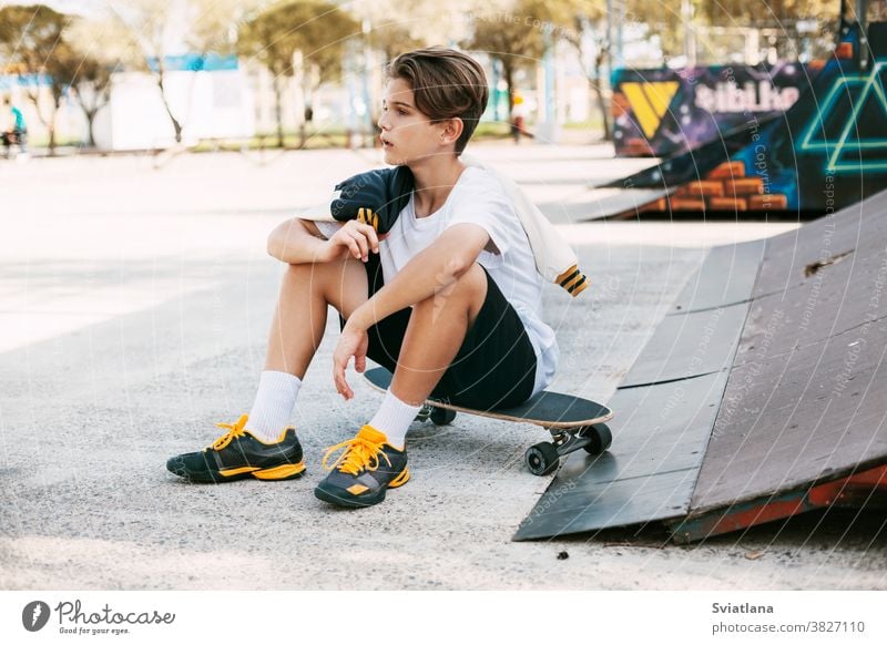 A beautiful teenager is sitting on a skateboard in a special area of the Park. A boy is resting after riding in a skatepark. Active rest in the fresh air kids