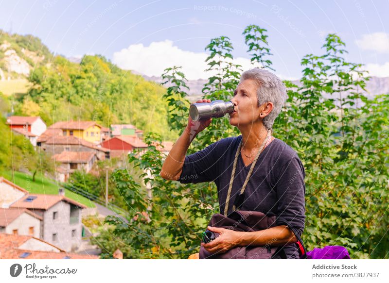 Mature female tourist drinking tea from thermos near greenery mountains traveler thirst nature highland house woman sky cloudy architecture old style hot drink