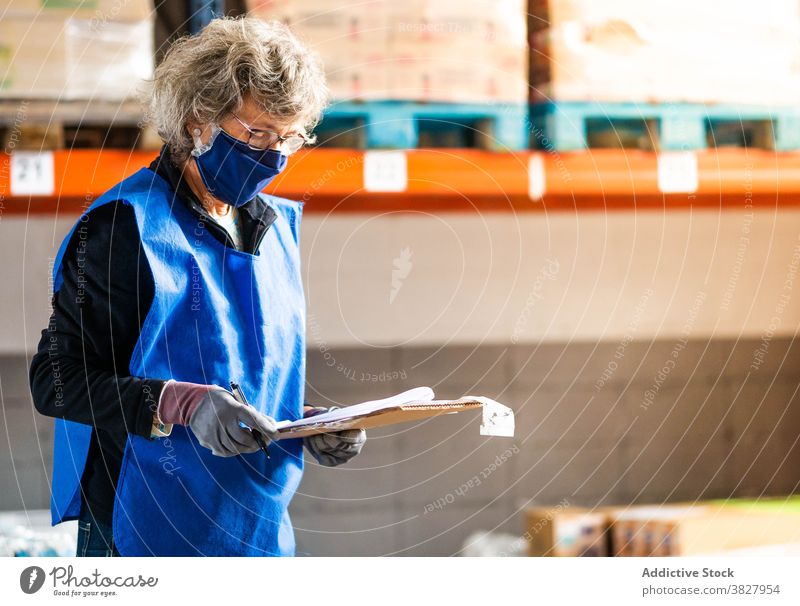 Mature female warehouse worker writing in clipboard near shelves woman record supervisor control paperwork take note write focus storehouse industrial at work
