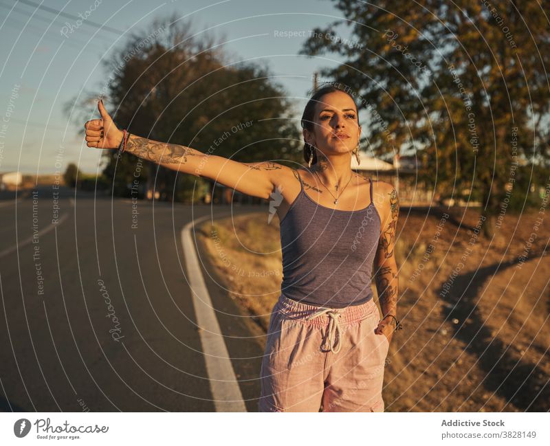 Woman catching car on road during vacation hitchhike travel woman outstretch summer traveler wait serious female sunset journey trip young stand style tattoo