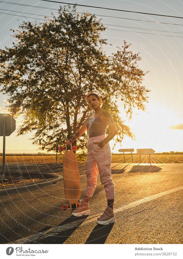Confident woman with longboard on road skater cool style sunset millennial individuality summer female young modern hipster contemporary stand carefree freedom