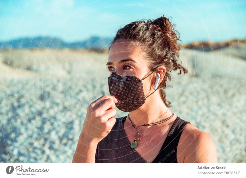 Sporty woman in mask and earphones standing among dry terrain sportswoman earbuds desert confident listen protect wireless strong determine coronavirus covid 19