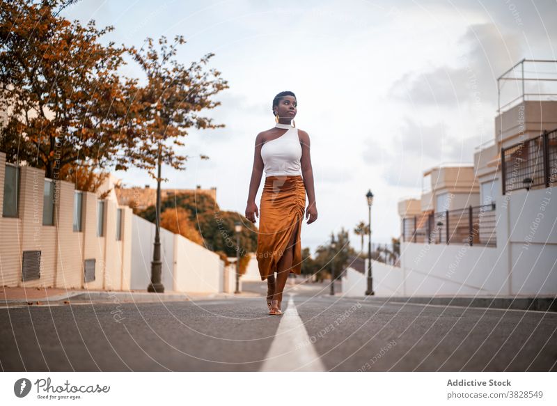 Stylish black woman walking on city road stylish apparel dreamy look building tropical earring contemplate nature sky modern style slim bright shabby rough