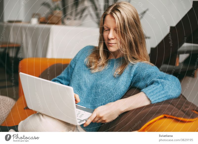 Young woman uses laptop, checks email news online sitting on sofa. Beautiful girl working on a computer, watching a webinar, studying at home young beautiful