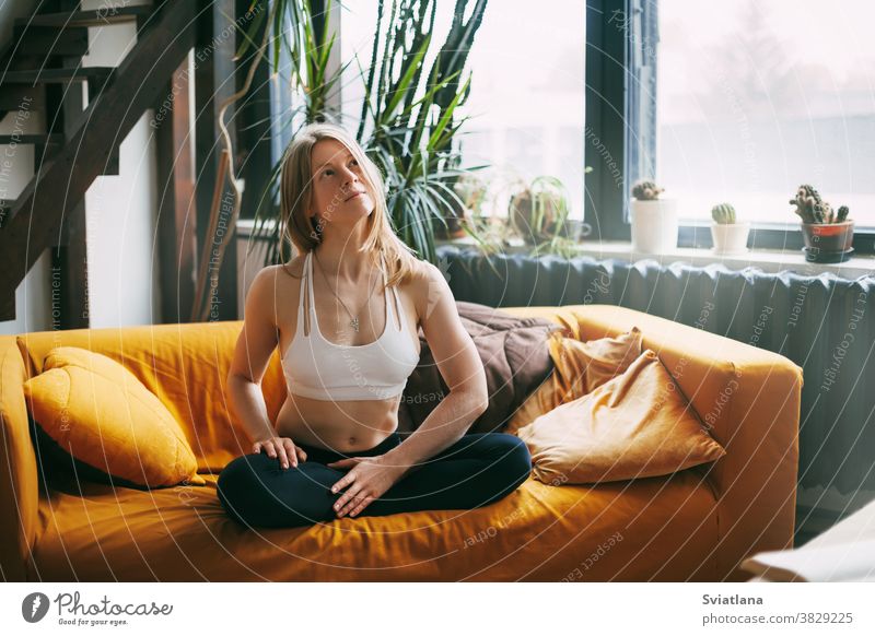 Serene calm girl sitting on the sofa in the Lotus position, doing yoga exercises for relaxation at home. The concept of peace of mind woman meditating couch